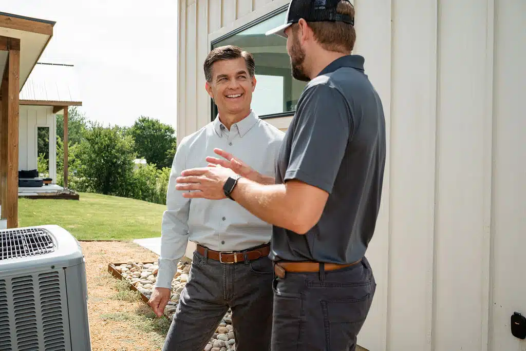 HVAC technician standing in front of an HVAC unit outside, speaking with a homeowner.