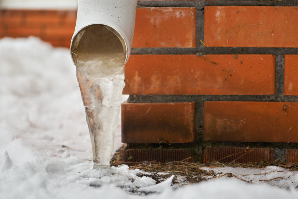 Drain pipe with frozen stream of water near brick wall of a cottage outdoors in winter